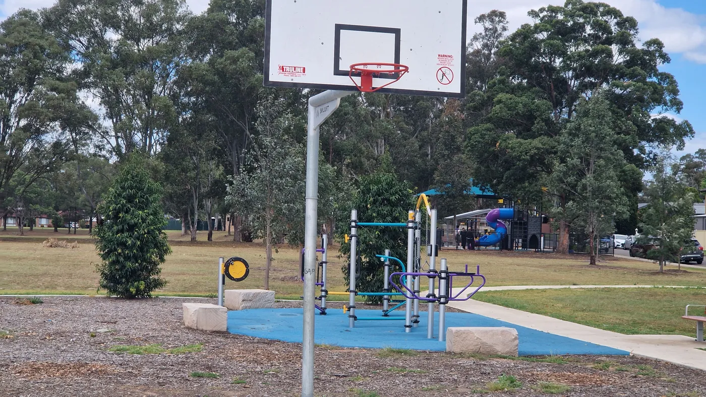 A colourful climbing frame on blue softfall beside a basketball hoop, with a fenced playground and spiral slide behind