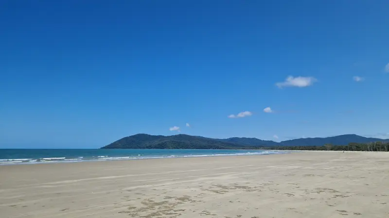 A long, empty sandy beach at low tide with forested headland and hills beyond